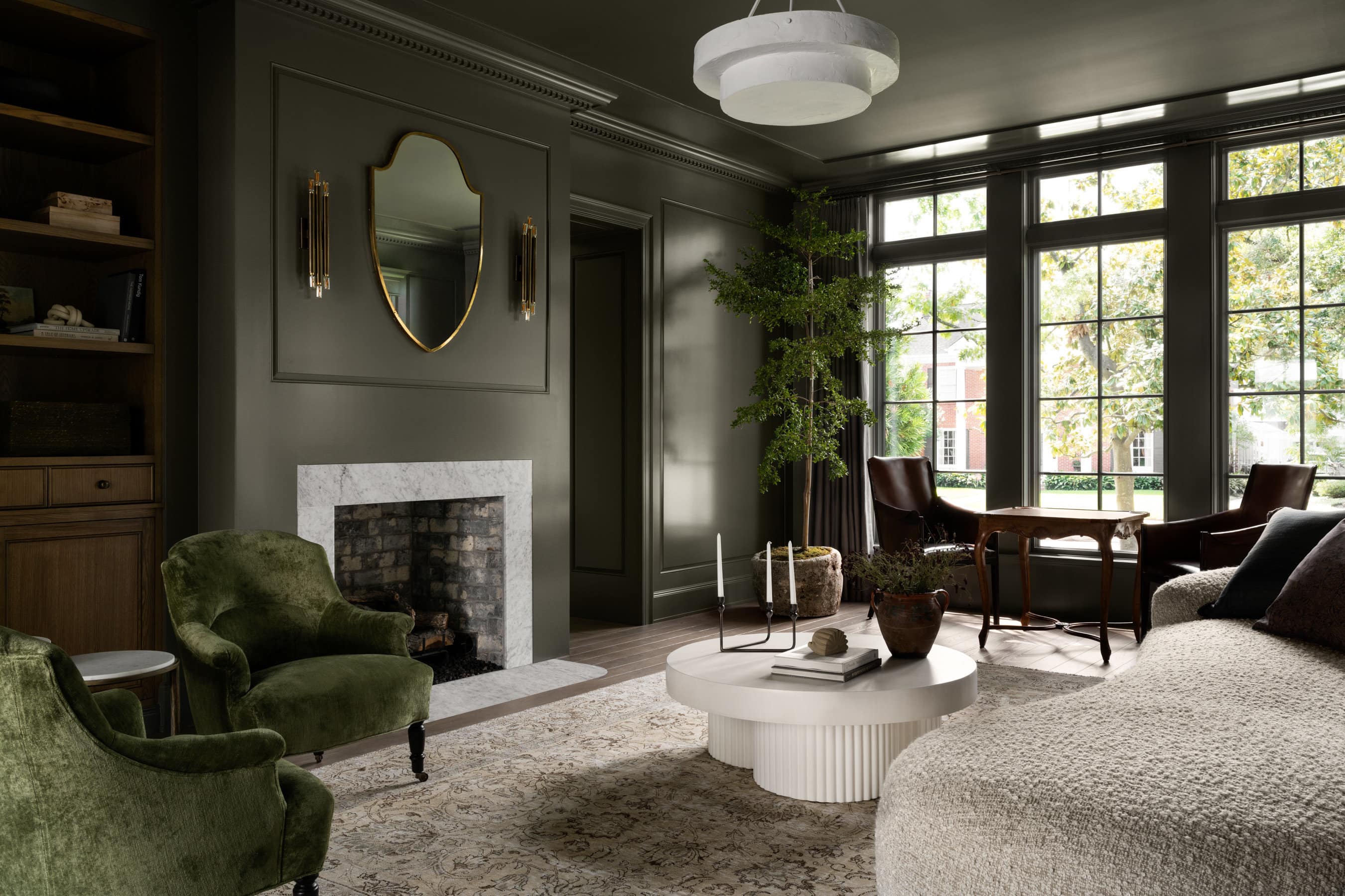 Elegant living room with dark green walls, large windows, a white marble fireplace, gold mirror, green velvet chair, white round coffee table, cozy sofa, armchairs, and a tall leafy plant by the window, by Studio McGee.