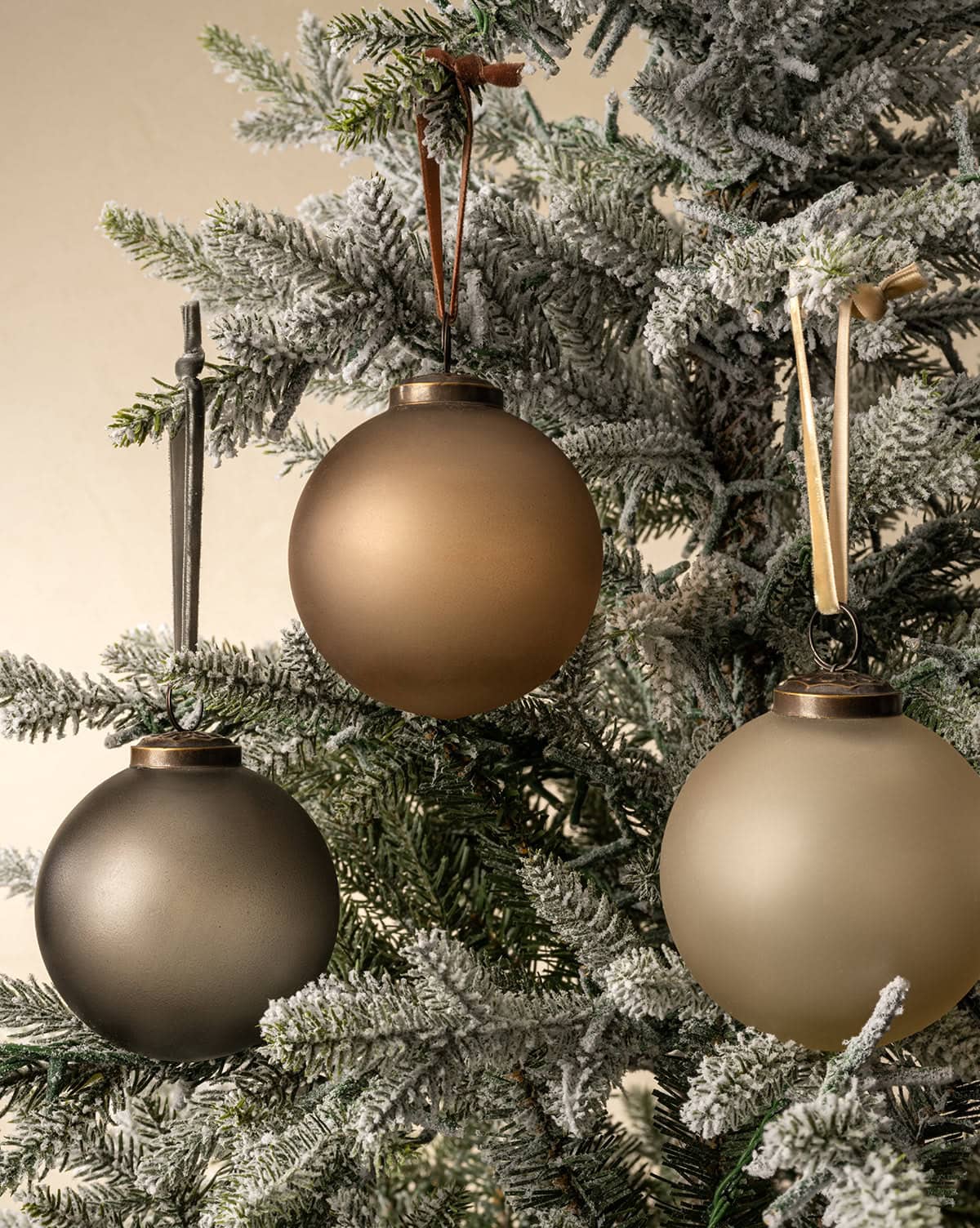 Neutral bulb ornaments designed in matte color finishes hang from velvet ties shown on a snow-dusted tree from Studio McGee.