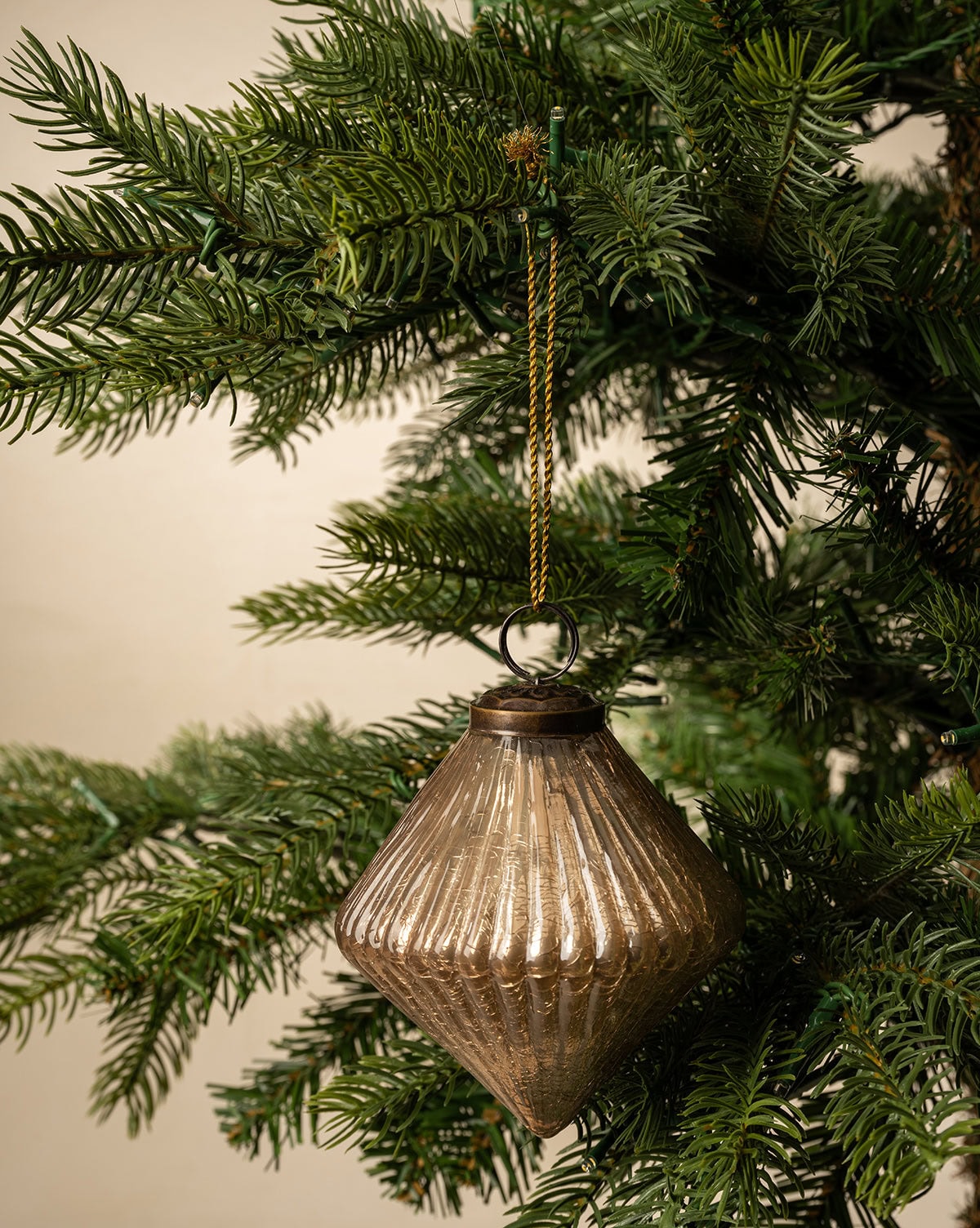 The Piper Ornament, is a gold diamond-shaped bulb shown hanging from a green Christmas tree branch from Studio McGee.