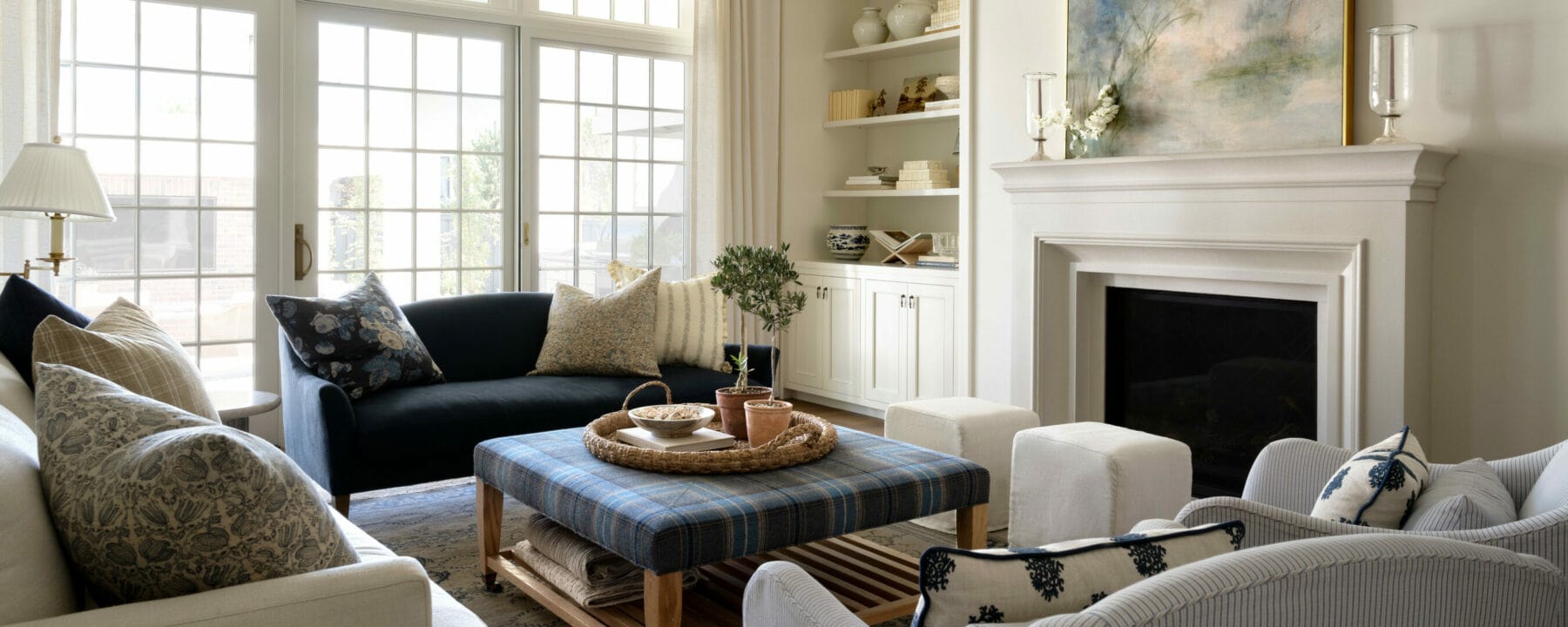 Bright living room with large windows, two sofas, armchairs, a blue plaid ottoman as a coffee table, built-in shelves, a fireplace with artwork above, and neutral, cozy decor with blue and beige accents, by Studio McGee.