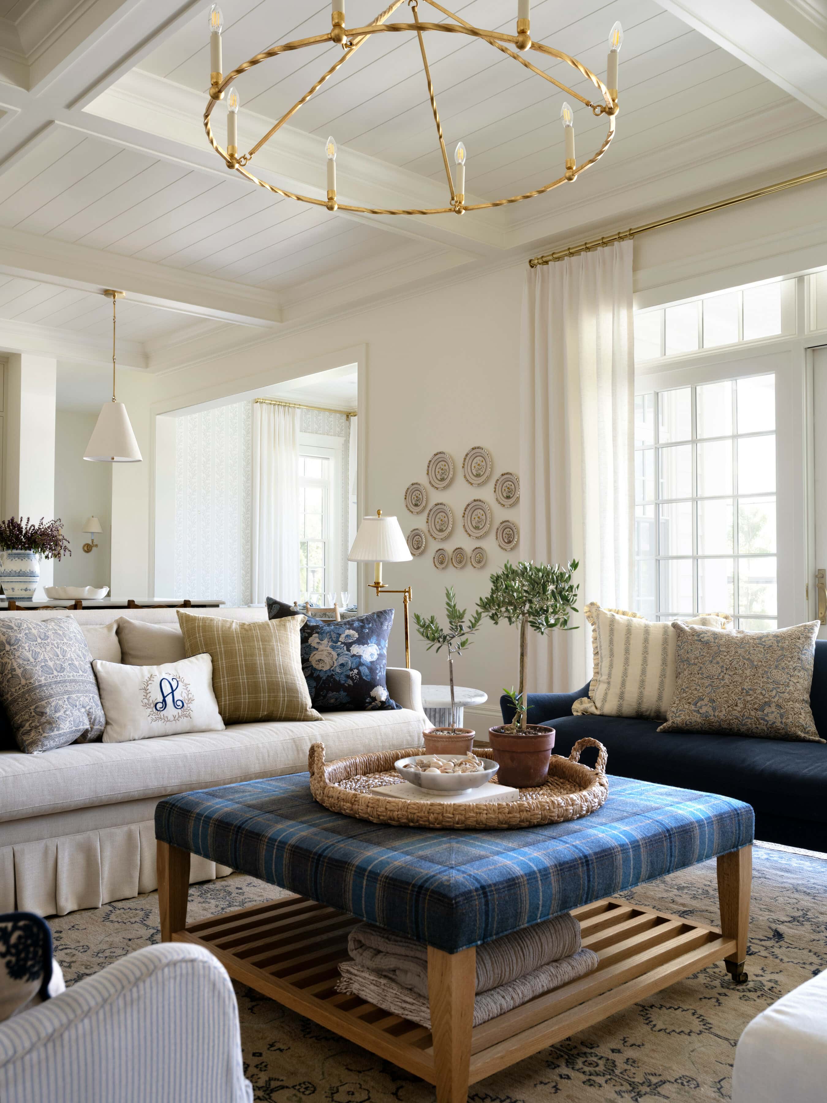 A bright, airy living room with light walls, a large window, beige sofas with patterned pillows, a blue plaid ottoman coffee table, a gold chandelier, and neutral decor accents, by Studio McGee.