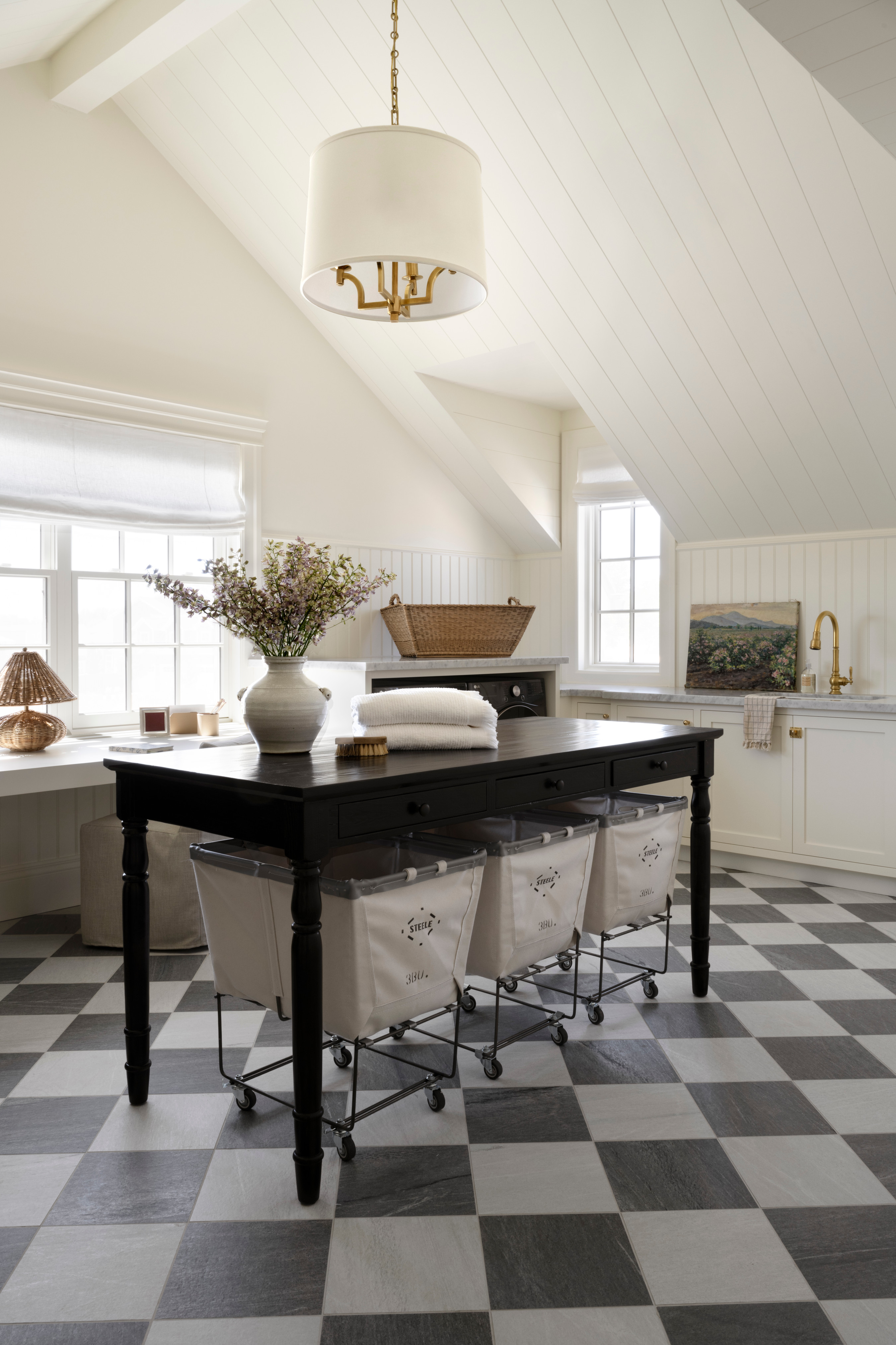 A well-organized laundry room with a black folding table, basket shelviong and cathedral ceiling with large pendant light from Studio McGee.