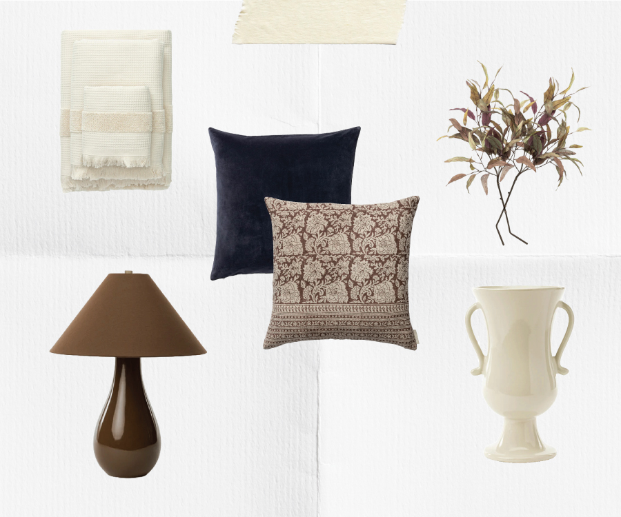 A collection of home decor items including a beige throw blanket, a cream rug, a faux leafy branch, a dark blue pillow, a patterned brown pillow, a brown table lamp, and a cream ceramic vase with handles, arranged on a white background, by McGee & Co.