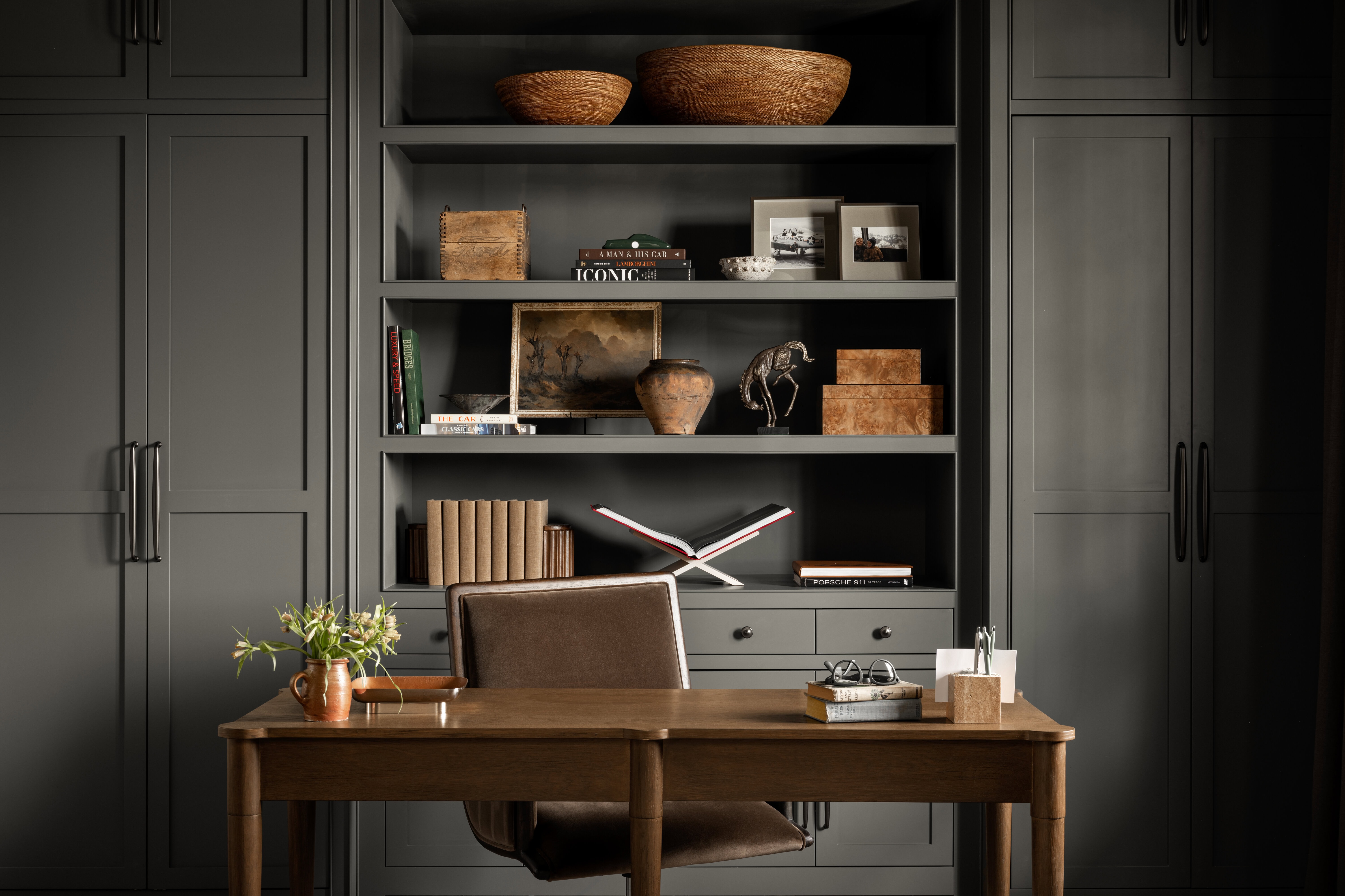 A stylish home office with a wooden desk, brown chair, and gray built-in shelves displaying books, baskets, ceramics, framed photos, sculptures, and decorative objects. A small plant sits on the desk, by McGee & Co.
