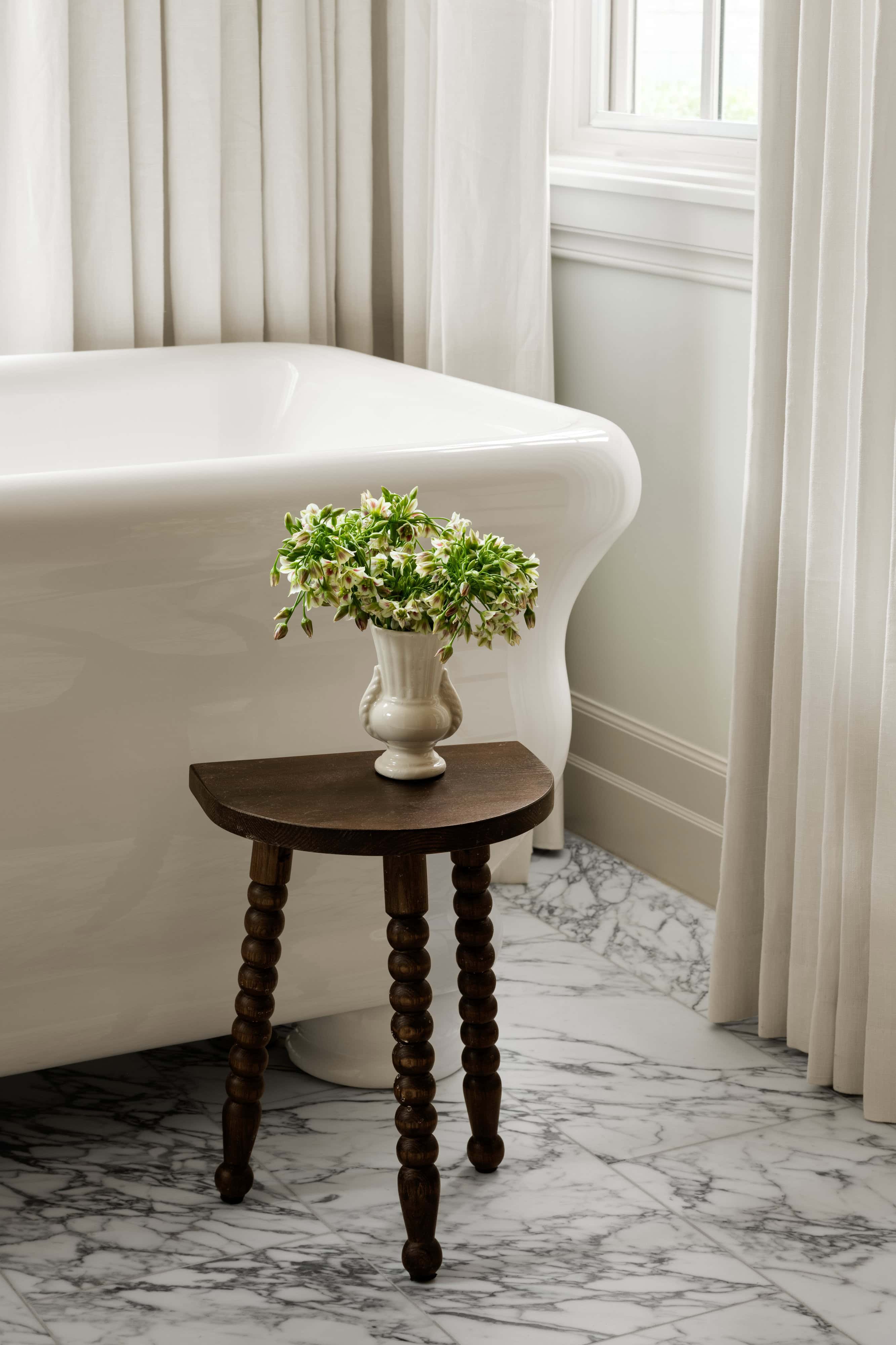A white bathtub and marble floor beside a window with white curtains; a small wooden table with a vase of green and white flowers is placed next to the tub, by McGee & Co.