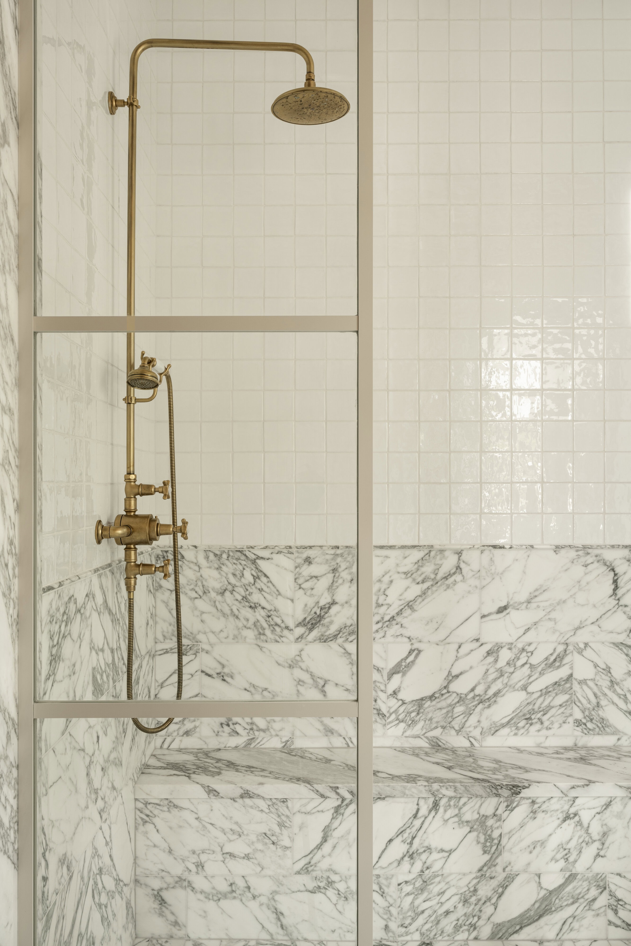A shower with brass fixtures is set against a wall of glossy white tiles and surrounded by white and gray marble surfaces, including a built-in bench, by McGee & Co.