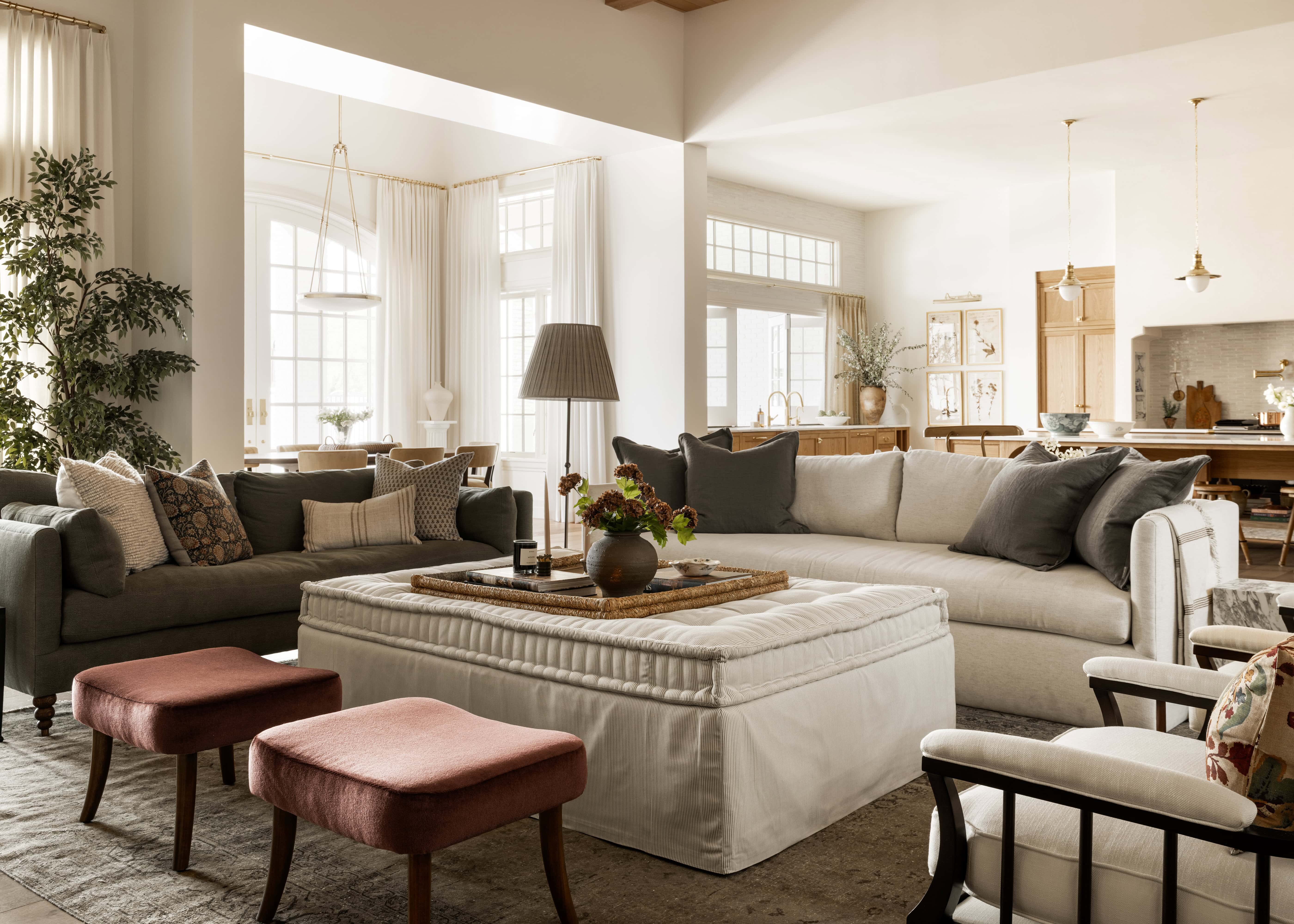 A spacious, sunlit living room with neutral-toned sofas, a large white ottoman, and red stools. There are decorative pillows, a table lamp, indoor plants, and a dining area visible in the background, by McGee & Co.