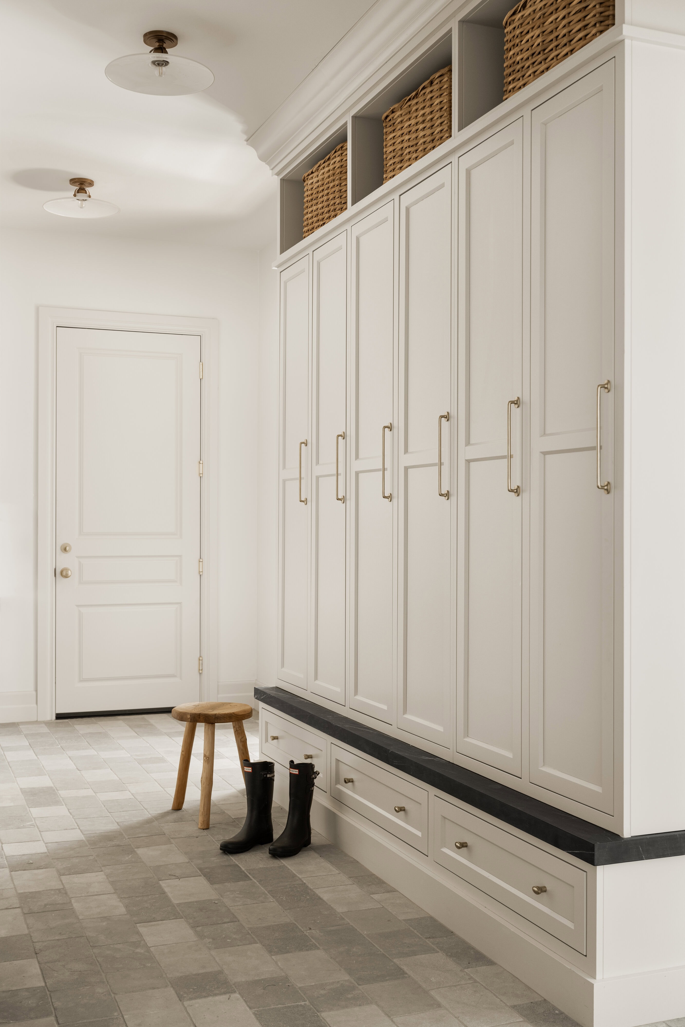 A bright mudroom with light cabinets, tall storage lockers with gold handles, baskets on top shelves, gray-tiled floor, a wooden stool, and black rain boots next to the bench. A white door is in the background, by McGee & Co.