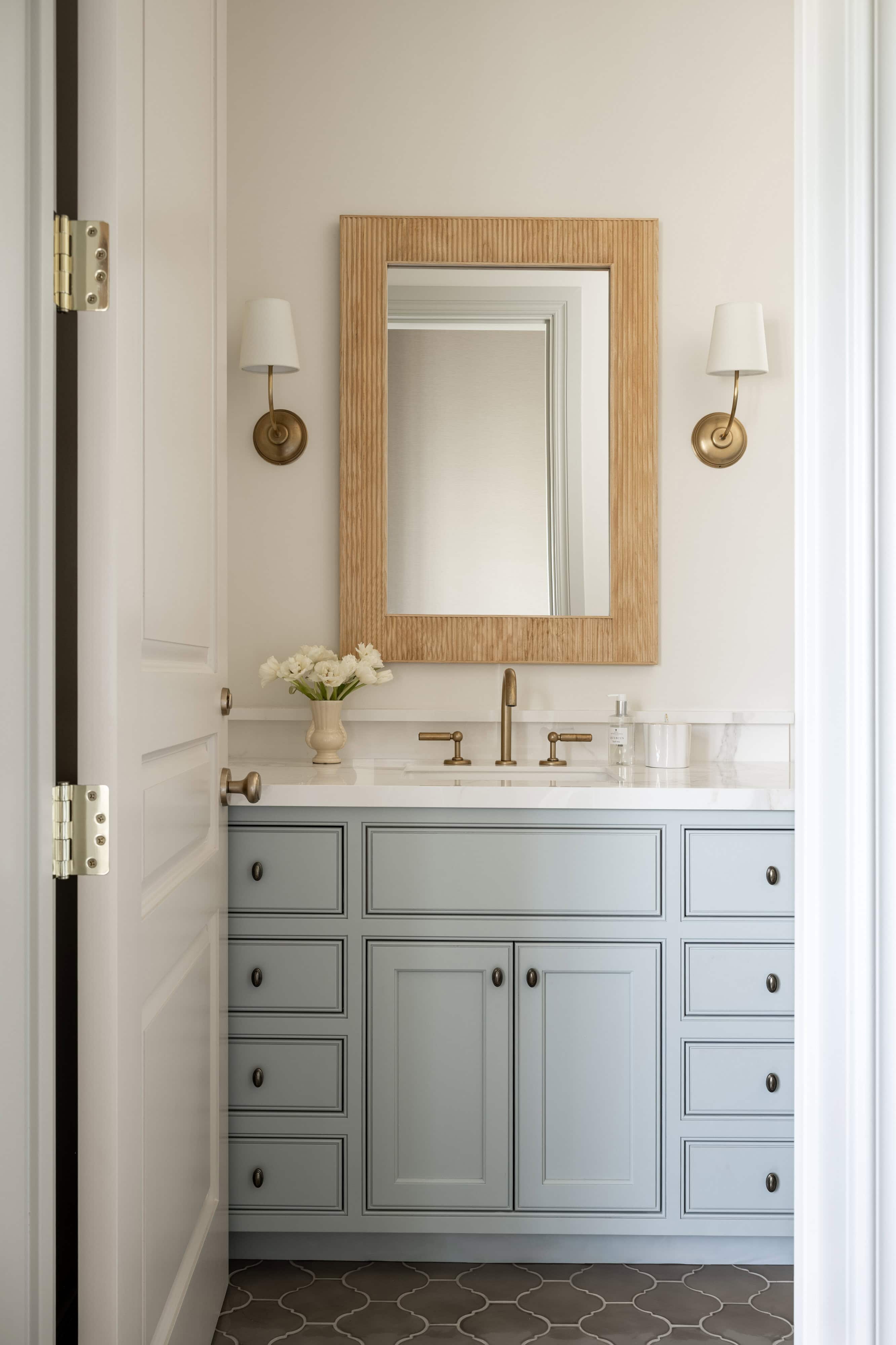 A small bathroom with light blue vanity cabinets, a white countertop, brass fixtures, a wood-framed mirror, two wall sconces, a vase of white flowers, and patterned dark tile flooring by McGee & Co.