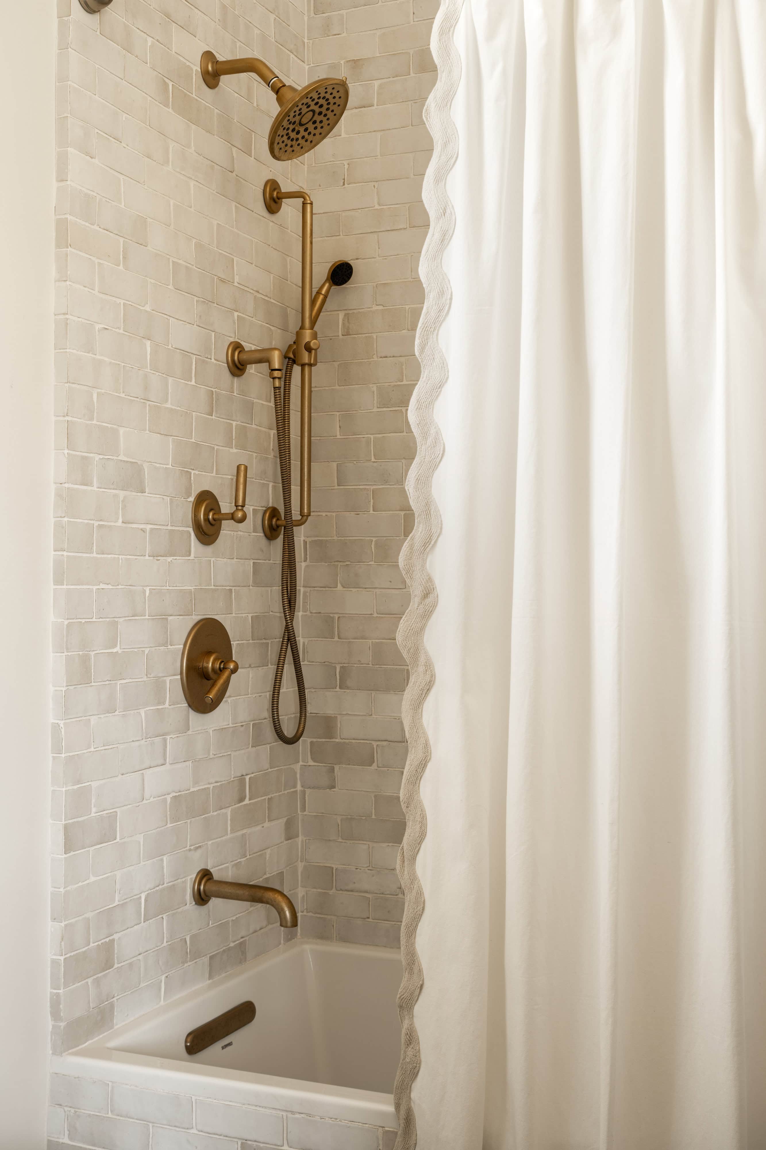 A modern bathtub and shower area with white subway tiles, brass fixtures including a rainfall showerhead and handheld sprayer, and a white shower curtain with a scalloped edge, by McGee & Co.