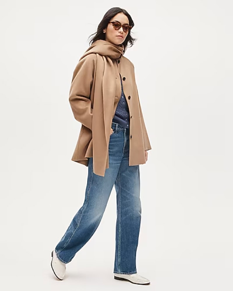 A woman wearing sunglasses, a tan hooded coat over a dark top, blue jeans, and white shoes walks confidently against a plain light background, by McGee & Co.