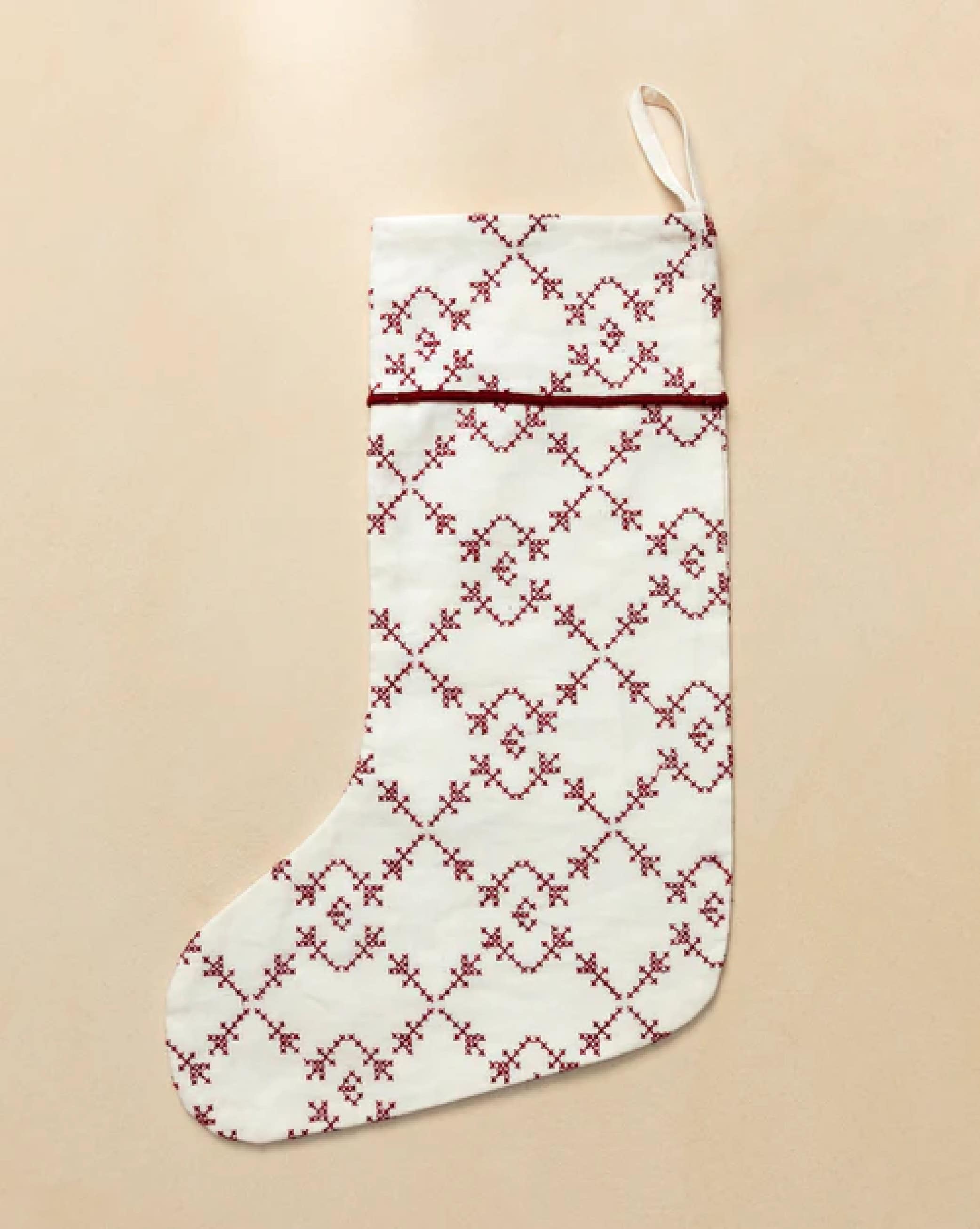 A white Christmas stocking with a red embroidered geometric pattern and a hanging loop, displayed on a beige background, by McGee & Co.