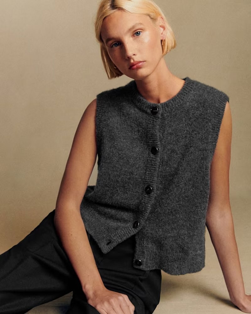 A woman with short blonde hair sits against a neutral background, wearing a sleeveless, dark gray button-up sweater vest and black pants, looking slightly to the side with a relaxed expression, by McGee & Co.