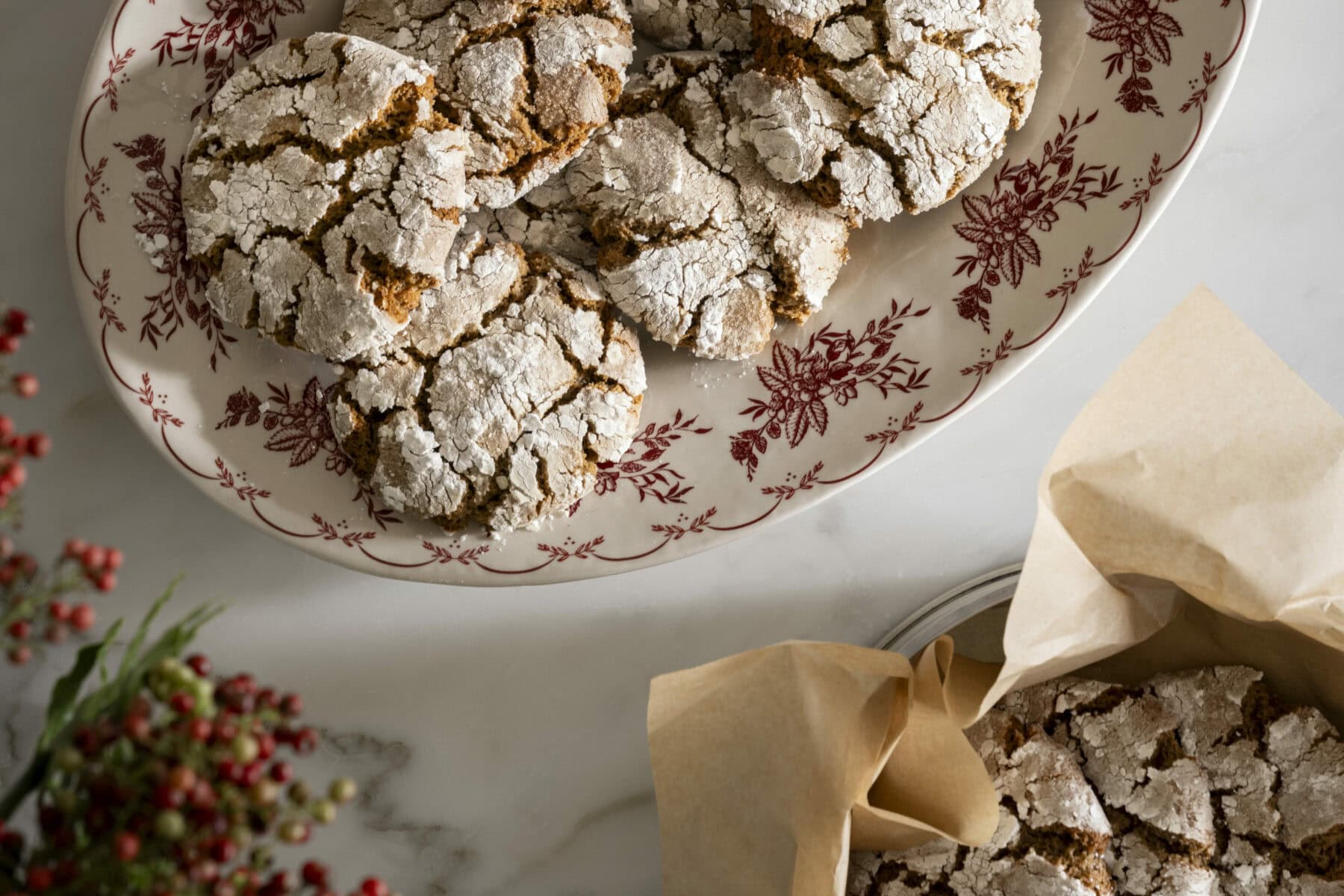 A decorative plate with powdered sugar-crusted cookies sits on a marble surface next to a parchment-lined tin with more cookies. Red berries are partially visible in the corner, by McGee & Co.
