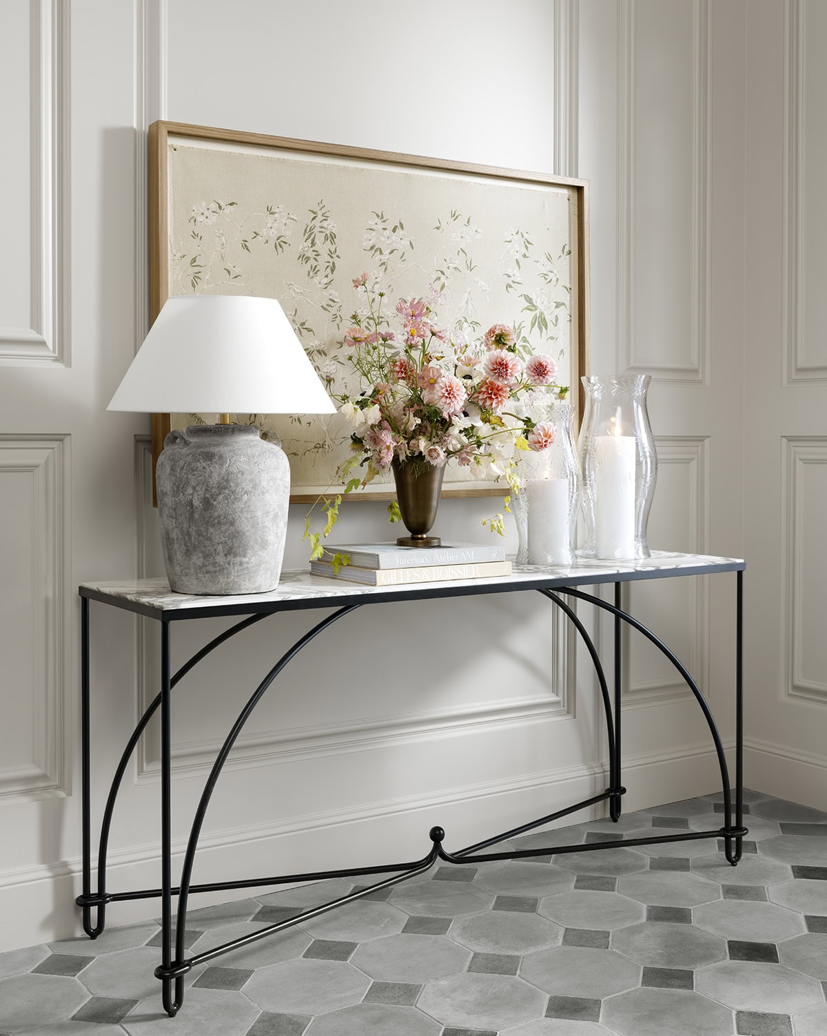 A marble-top console table with a gray lamp, a vase of pink and orange flowers, books, candles, and glass vases stands against a white paneled wall, beneath a framed botanical artwork on hexagon tile flooring, by McGee & Co.