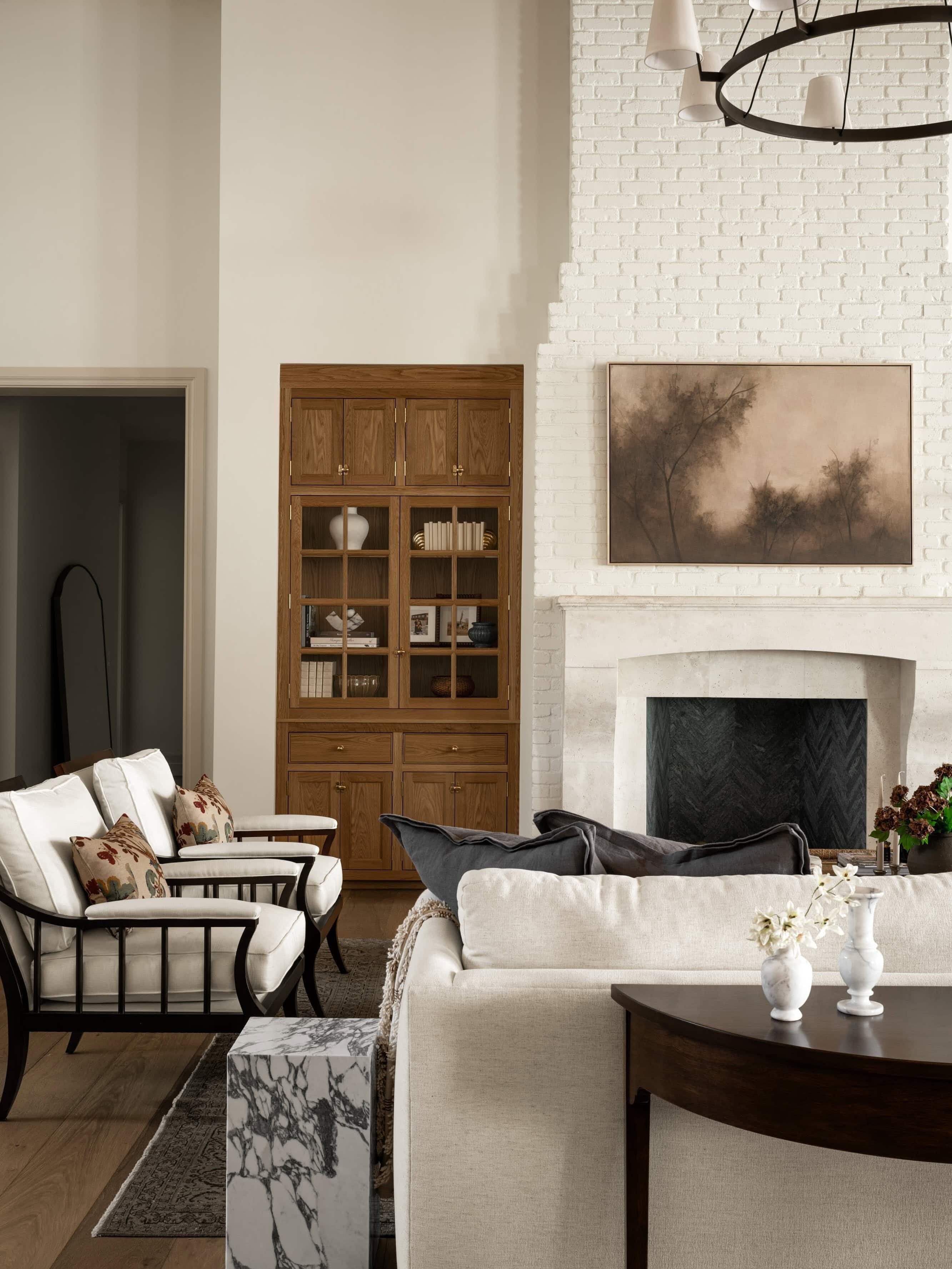 A cozy living room with cream-colored seating, a white brick fireplace, wooden cabinetry filled with books, and a nature-themed painting above the mantel. Neutral tones and soft lighting create a serene atmosphere, by McGee & Co.