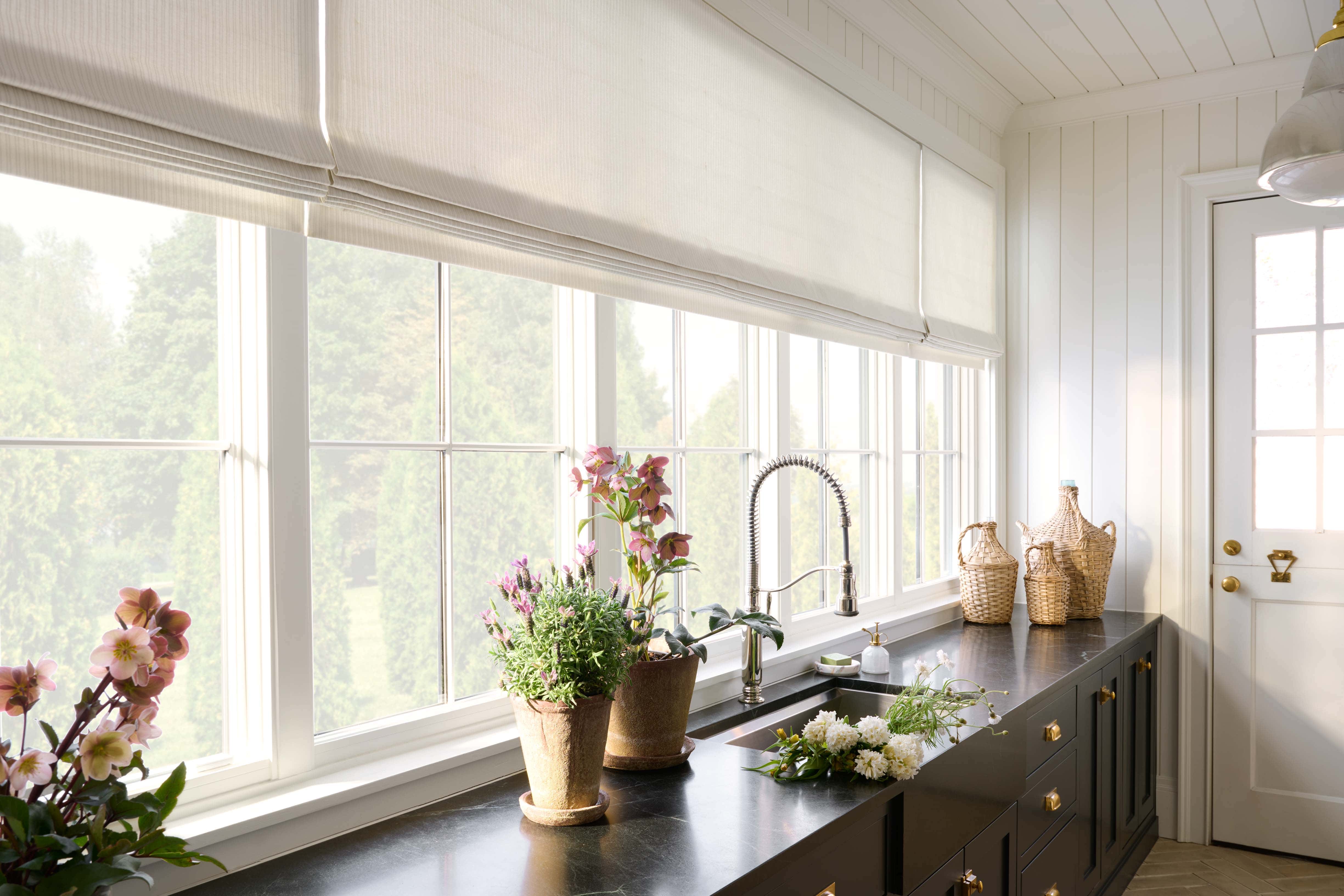 A bright kitchen with large windows, white roman shades, black countertops, a stainless steel sink, potted flowers, wicker baskets, and a bouquet of white flowers on the counter. Natural light fills the room, by McGee & Co.
