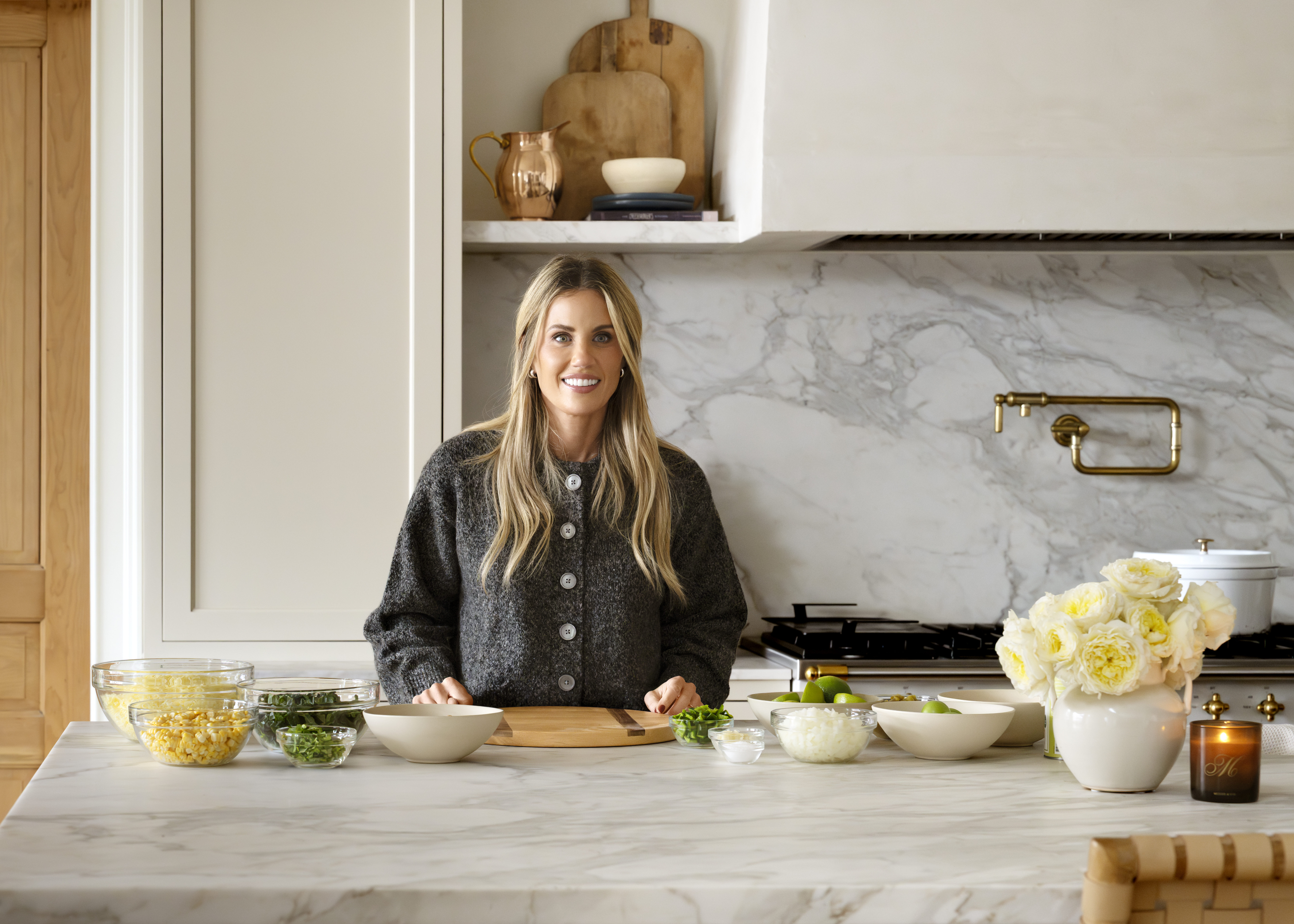 A woman stands and smiles in a modern kitchen, preparing food on a marble countertop with bowls of chopped vegetables, herbs, pasta, and a vase of white roses beside her, by McGee & Co.
