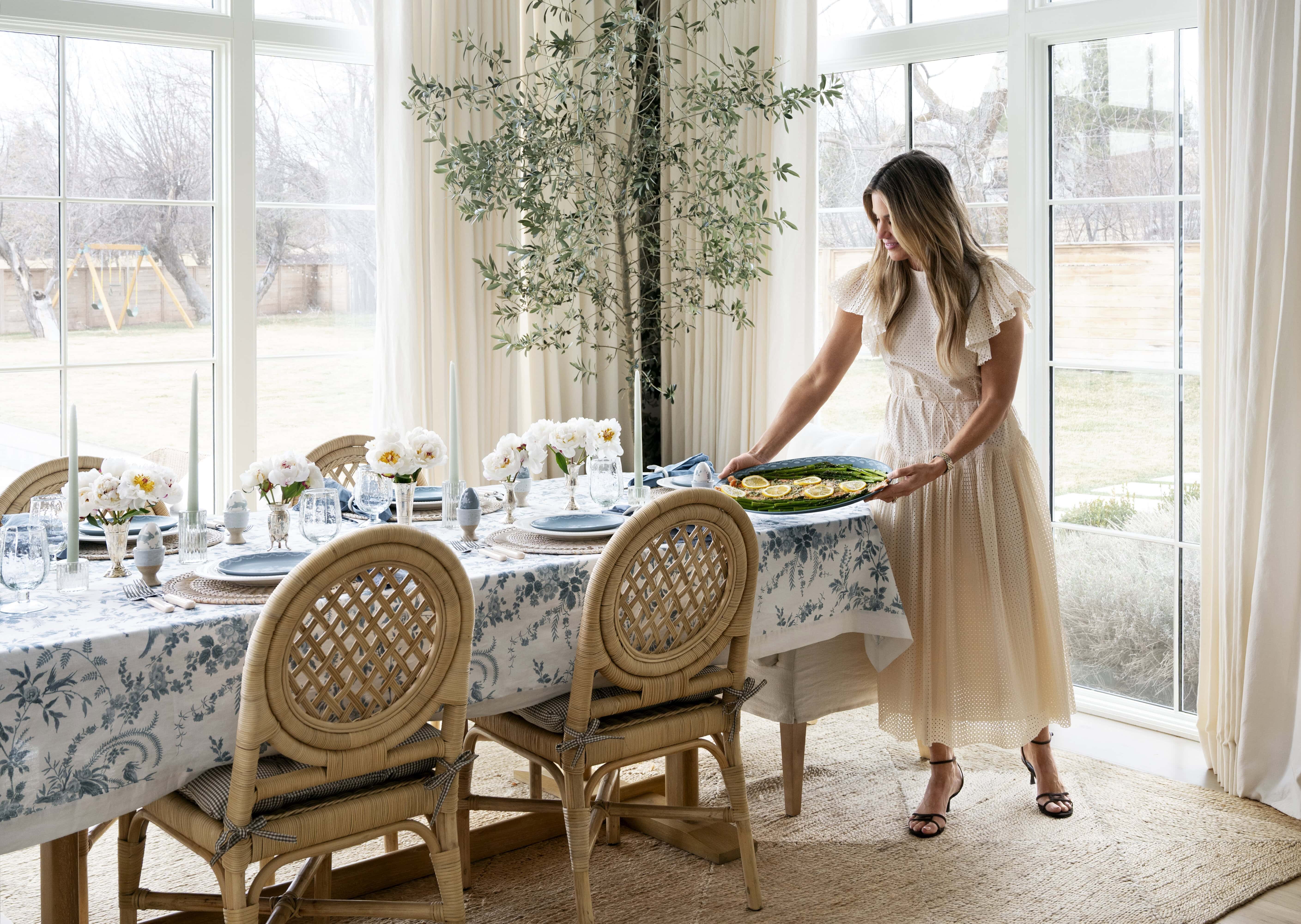 A woman in a light dress sets a platter of food on an elegantly set dining table decorated with white flowers and patterned tablecloth, in a bright room with large windows and an indoor tree, by McGee & Co.