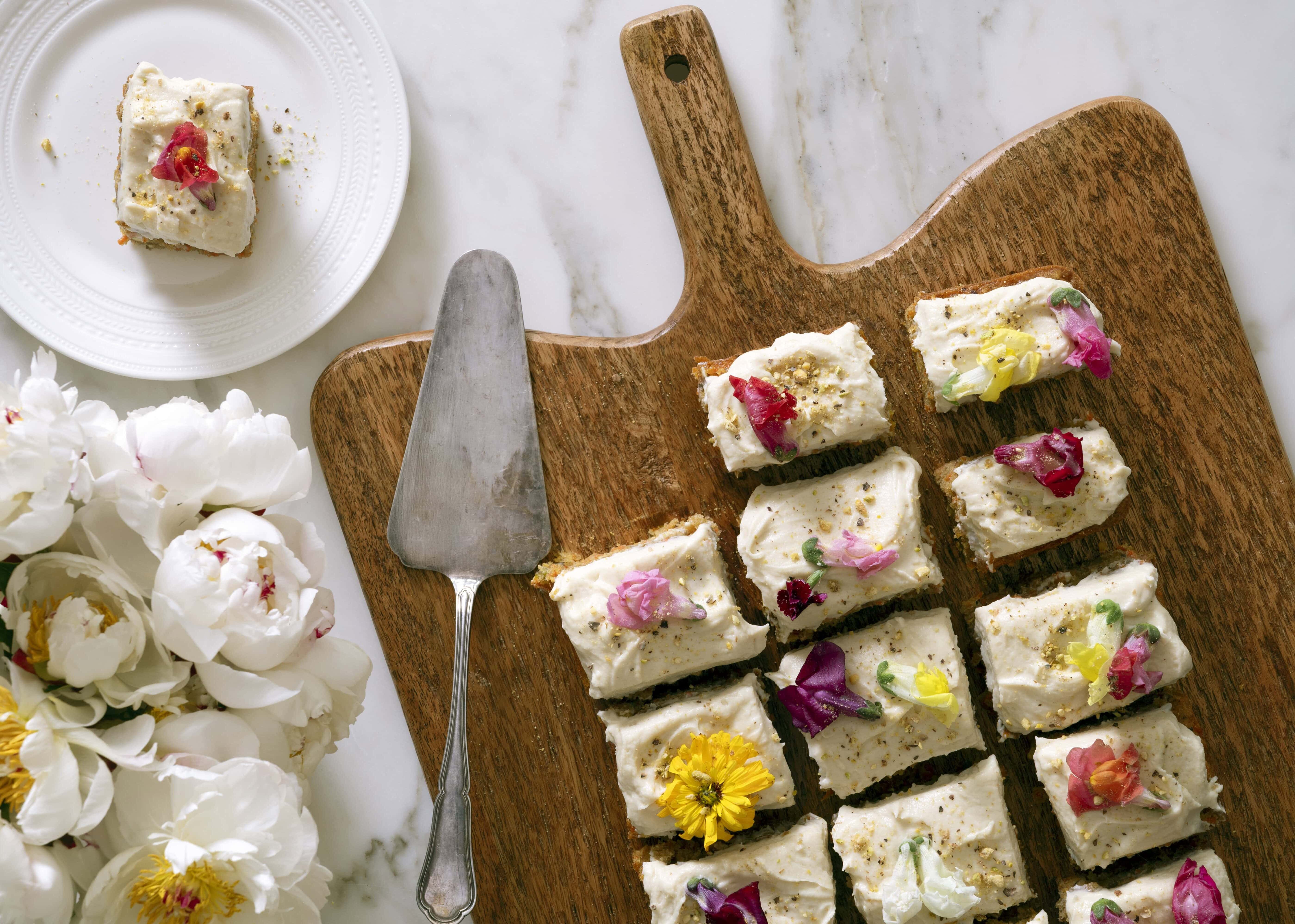 Sliced frosted cake squares topped with colorful edible flowers on a wooden board, with one piece on a small white plate nearby. A cake server and a bunch of white flowers are also visible on a marble surface, by McGee & Co.
