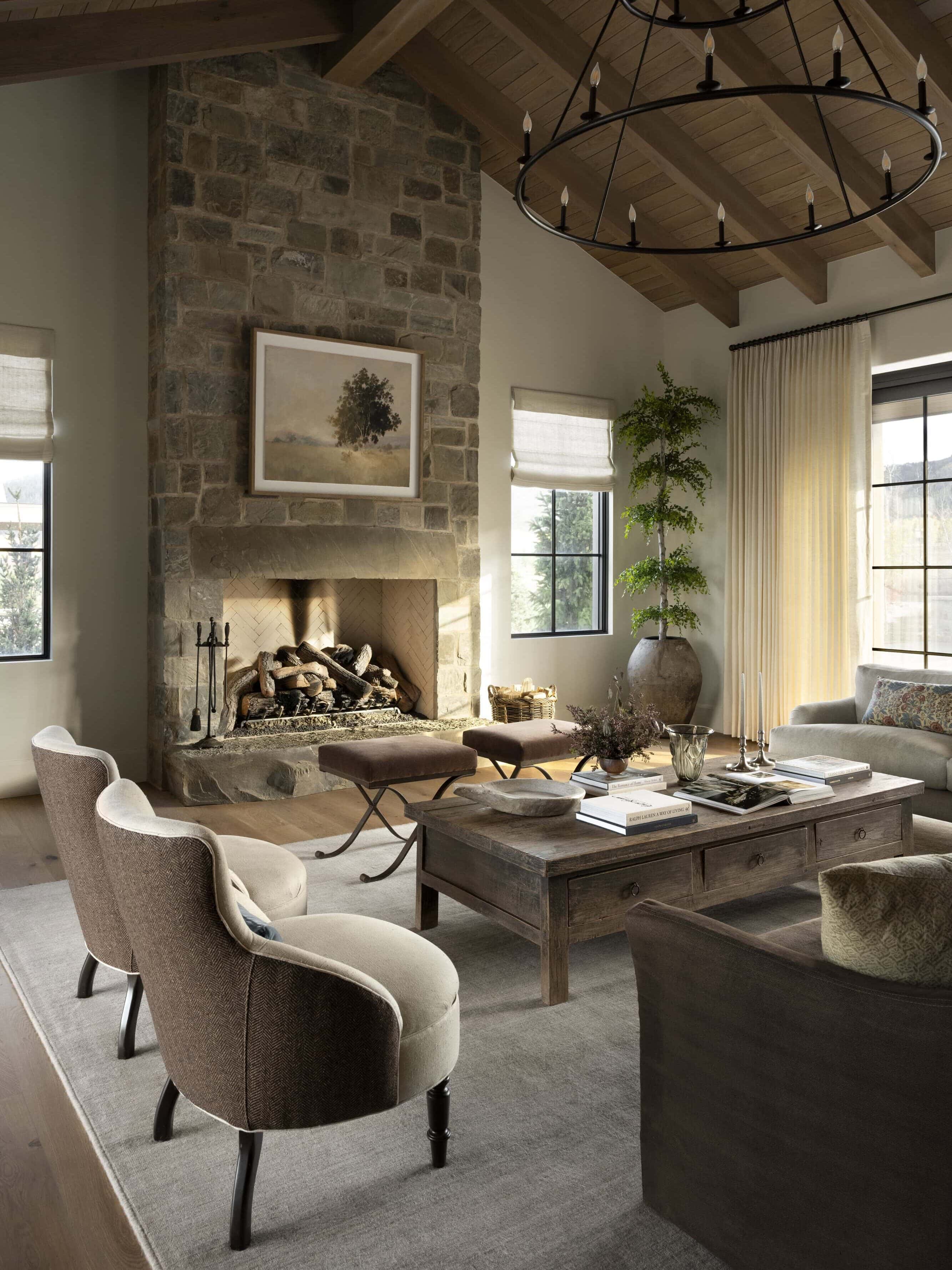 A cozy living room with a stone fireplace, neutral-toned furniture, a wooden coffee table, a chandelier, large windows with natural light, and a tall potted plant in the corner, by McGee & Co.