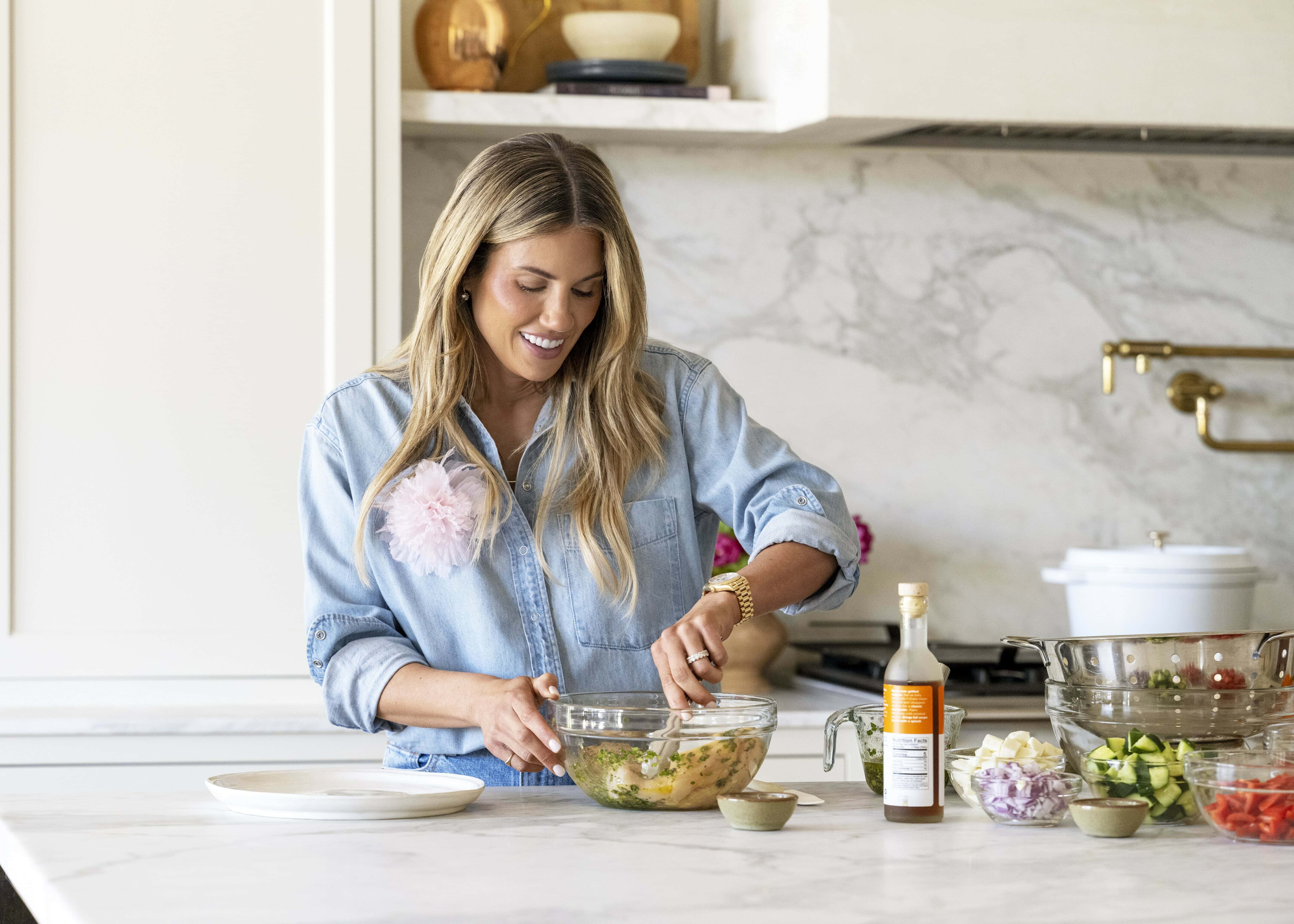 A woman in a denim shirt smiles while mixing ingredients in a glass bowl in a modern kitchen, with vegetables and condiments on the marble countertop around her, by McGee & Co.